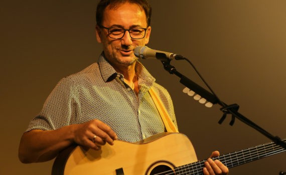 Lutz Scheufler lächelnd mit Gitarre am Mikrofon